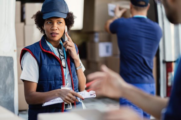 Black female deliverer communicating with coworker while talking on mobile phone and organizing package delivery.