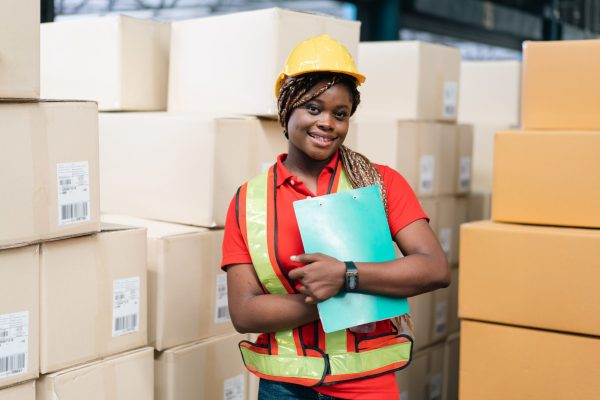 Portrait of smiling African female worker holding checklist report document with parcel boxes in the background looking at camera. Warehouse woman working for logistic business